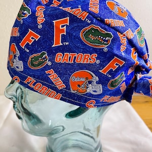 May include: Blue scrub cap with an all-over pattern featuring the University of Florida Gators logo, including the letter "F", a Gator head, and a football helmet. The words "Gators" and "Florida Pride" are also visible.