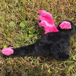 May include: A black and pink plush toy shaped like a dragon with wings and a long tail. The toy is lying on a bed of green grass.