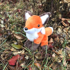 May include: A soft toy fox in orange, white, brown, and black. The plush fox has a white chest and tail tip, brown paws and ears, and is positioned amongst grass and fallen leaves.
