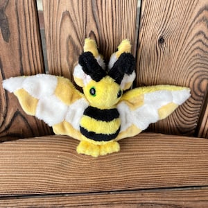 May include: A yellow and black bee plush toy with white wings. The bee has large green eyes and a fuzzy texture.