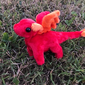 May include: A plush red dragon toy with orange wings and accents, set against a backdrop of green grass. The dragon has black eyes and a long tail, creating a whimsical and playful appearance.