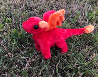 May include: A plush red dragon toy with orange wings and accents, set against a backdrop of green grass. The dragon has black eyes and a long tail, creating a whimsical and playful appearance.