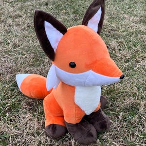 May include: A plush orange fox toy with brown ears and a white chest. The fox is sitting on a bed of brown grass.