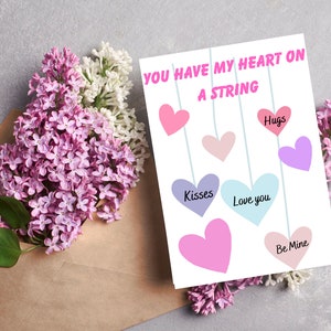 以下が含まれることがあります： 「You have my heart on a string」という文字が書かれた白いカードと、カラフルなハートが紐にぶら下がっています。ハートには「Hugs」「Love you」「Kisses」「Be Mine」の文字が書かれています。