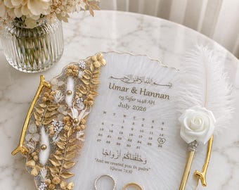May include: A white resin tray with gold handles, decorated with dried flowers and gold leaves. It features the text Umar & Hannan, July 2026, and Quran 78:8. Two rings rest on the surface next to a white feather pen with a white rose accent.