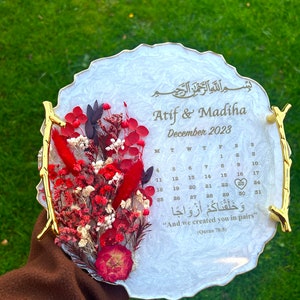 Pode incluir: Bandeja de resina branca com borda dourada e um calendário para dezembro de 2023. A bandeja tem um arranjo floral com flores secas vermelhas e brancas. O texto "Atif & Madiha" e um versículo do Corão em árabe estão inscritos na bandeja.