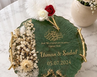 May include: A round green resin tray with gold branch-style handles, decorated with dried white flowers, a white and red rose, and white feathers. It features gold calligraphy, the text "And We created you in pairs (Quran 78:8)", "Usman & Sadaf", and "05.07.2024".