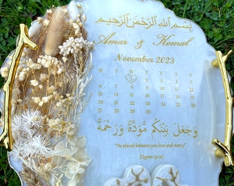 May include: A white resin ring dish with a gold trim and dried flowers. The dish features a calendar for November 2023 with a heart highlighted on the 8th. It also includes the Arabic text "بسْمِ اللَّهِ الرَّحْمَنِ الرَّحِيمِ" and the English translation "He placed between you love and mercy" (Quran 30:21).