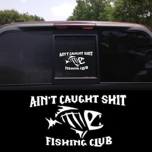 May include: A white vinyl decal on a car window with the text "AIN'T CAUGHT SHIT" above a fish skeleton graphic and the words "FISHING CLUB" below. The decal is displayed on a black background.