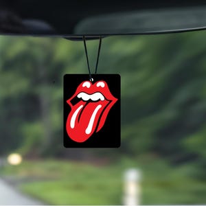 Könnte beinhalten: Ein schwarzer, rechteckiger Lufterfrischer mit dem ikonischen roten Lippen- und Zungen-Logo, der am Rückspiegel eines Autos hängt. Das Logo ist ein auffälliges Grafikdesign auf schwarzem Hintergrund, mit verschwommenem grünem Hintergrund.