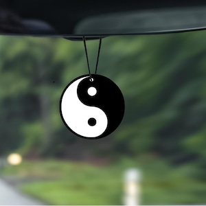 May include: A black and white Yin and Yang symbol car air freshener hanging from a rearview mirror. The air freshener is round with a black and white design. The background is a blurred view of a road and trees.