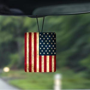 May include: Rectangular air freshener with the American flag design. The flag has red and white stripes and a blue field with white stars. The air freshener is suspended from a rearview mirror, against a blurred green backdrop.