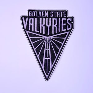 May include: A triangular embroidered patch featuring a black background with light purple stitching. The text reads GOLDEN STATE VALKYRIES in a bold font, positioned above a stylized graphic of a road converging toward a central point.
