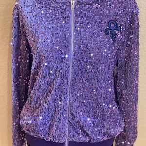 May include: A purple sequined bomber jacket with a purple velvet lining and a zipper closure. The jacket features a purple and blue embroidered symbol on the left chest.
