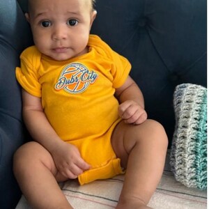 Puede incluir: Un bebé con un mono amarillo con un gráfico de baloncesto y el texto "Dubs City" está sentado en una manta a rayas. El bebé tiene el pelo castaño y los ojos marrones.