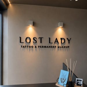 May include: A beige wall features black 3D lettering that reads LOST LADY TATTOO & PERMANENT MAKEUP, illuminated by two square wall-mounted lights. Below, a blue card with a line drawing and a small black and white photo sit on wooden stands.