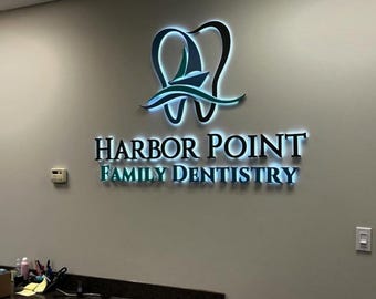 May include: Illuminated sign for Harbor Point Family Dentistry. The sign features a stylized tooth and wave design in teal and white, with the practice name in black and teal lettering. The sign is mounted on a light gray wall.