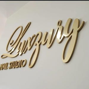 Puede incluir: Un letrero de pared tridimensional que presenta la palabra Luxury en una fuente cursiva grande de color dorado, colocada sobre las palabras NAIL STUDIO en una fuente sans-serif dorada más pequeña, todo montado en una pared blanca.