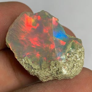 May include: A raw, uncut opal gemstone featuring a translucent, milky body with vibrant flashes of neon red and electric blue play-of-colour. The stone has a jagged, irregular shape with a rough, textured, light-coloured mineral matrix base.