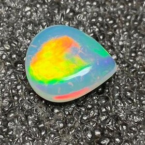 Może przedstawiać: Opal w kształcie łzy z żywą paletą barw. Opal ma półprzezroczysty korpus z wewnętrznymi błyskami żółtego, pomarańczowego, zielonego i czerwonego. Kamień osadzony jest na teksturowanej, ciemnoszarej powierzchni.