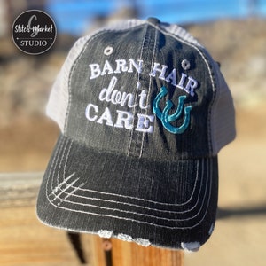 May include: A black and gray distressed trucker hat with a white mesh back. The hat has white embroidery that reads "Barn Hair don't care" with two turquoise horseshoes.