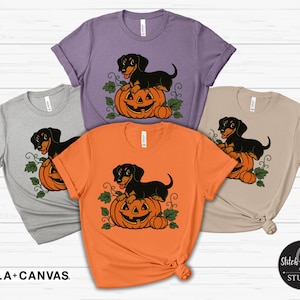 May include: A selection of Bella+Canvas t-shirts in different colours, each displaying a black dachshund dog perched on an orange pumpkin. These Halloween-themed shirts feature the text "BELLA+CANVAS".
