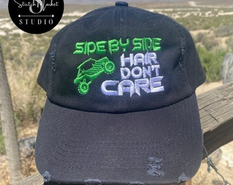 May include: Black baseball cap with distressed detailing. The cap features white and green embroidery that reads "Side by Side, Hair Don't Care".
