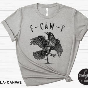 May include: A light gray Bella+Canvas t-shirt featuring a black graphic of a crow with outstretched wings and the text "F-CAW-F". The crow is depicted in a detailed, artistic style. The shirt has rolled sleeves.
