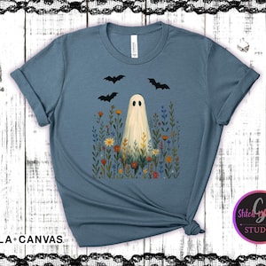 May include: A blue Bella+Canvas t-shirt featuring a whimsical Halloween design. The graphic includes a ghost in a field of flowers with three bats flying above. The shirt has rolled-up sleeves and a knotted hem.