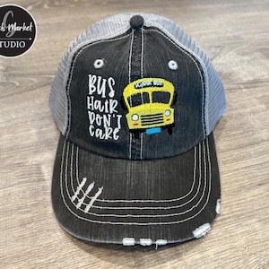 May include: A distressed black trucker hat with a grey mesh back. The front features the text "BUS HAIR DON'T CARE" and a yellow school bus graphic. The hat has a curved brim and white stitching.