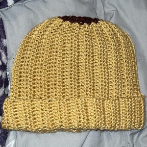 May include: A handmade, mustard yellow crochet beanie with a folded brim. The top of the hat features a dark brown crocheted accent. The hat is made with a ribbed stitch pattern.