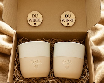 May include: A cardboard gift box containing two two-toned ceramic mugs with a speckled white top and a solid beige bottom. The mugs are engraved with the words OMA and OPA above a heart. Two wooden discs engraved with DU WIRST and a heart are included.