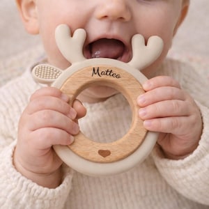 Könnte beinhalten: Ein runder Beißring mit einem hellbeigen Silikonrahmen in Form eines Hirschgeweihs und einem glatten Holzkern. Der Holzring ist mit dem Namen Matteo in Schreibschrift sowie einem kleinen Herzsymbol am unteren Rand graviert.