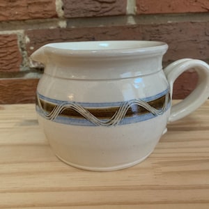 May include: A white ceramic pitcher with a brown and blue wavy design. The pitcher has a handle and a spout.