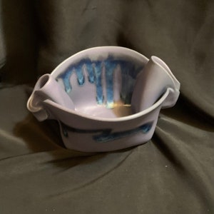 May include: A ceramic bowl with two handles and a blue and purple glaze. The bowl has a unique, abstract design.