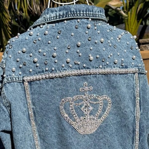May include: A light-wash denim jacket adorned with pearls and rhinestones. The back showcases a rhinestone-outlined crown design. Pearls are scattered across the shoulders and sleeves, with rhinestone trim outlining the jacket's seams.