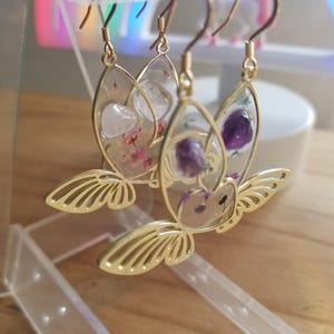 Könnte beinhalten: Goldene Schmetterlings-Ohrringe mit klarem Harz und gepressten Blumen und Kristallen. Die Ohrringe hängen an einem Ausstellungsständer.