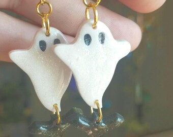 Op de afbeelding: Een paar oorbelletjes met witte spookbedels met zwarte ogen en gouden accenten. De spoken zijn bevestigd aan zwarte vleermuisbedels met gouden hoepels.
