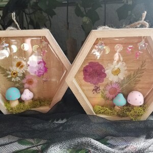 May include: Two hexagonal wooden frames with clear resin inserts. Each frame contains pressed flowers and a pair of miniature mushrooms. The mushrooms are painted in pastel colors with white polka dots. The frames are decorated with gold stars and moons.
