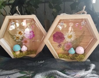 May include: Two hexagonal wooden frames with clear resin inserts. Each frame contains pressed flowers and a pair of miniature mushrooms. The mushrooms are painted in pastel colours with white polka dots. The frames are decorated with gold stars and moons.