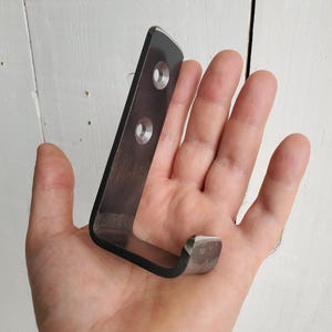 Op de afbeelding: Een zwarte metalen haak met twee gaten voor montage. De haak is gebogen in een hoek van 90 graden en heeft een afgeronde rand.