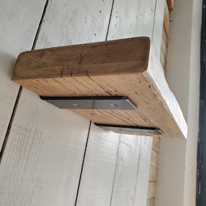 Pode incluir: Uma prateleira de madeira rústica com uma borda viva e um acabamento natural. A prateleira é suportada por dois suportes de metal preto. A prateleira está montada em uma parede branca.