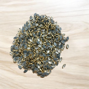 Può includere: Un mucchio di piccole perline ovali con un bordo dorato metallico e un centro grigio scuro riflettente. Le perline sono sparse su una superficie di legno chiaro, creando un aspetto strutturato.