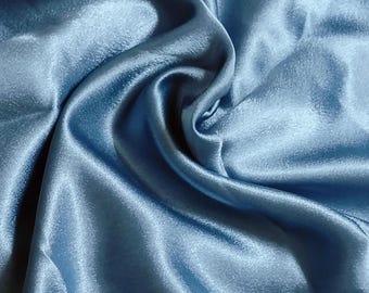 May include: A light blue fabric with a smooth, lustrous finish and a reflective surface. The material is gathered into soft, flowing folds and deep ripples, creating a dynamic texture that highlights the sheen of the light blue textile.