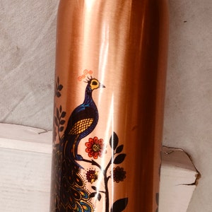 May include: A cylindrical copper water bottle with a metallic finish, featuring a printed graphic of a peacock with blue, yellow, and orange plumage perched on a branch with red and orange flowers. The bottle has a matching screw-top lid.