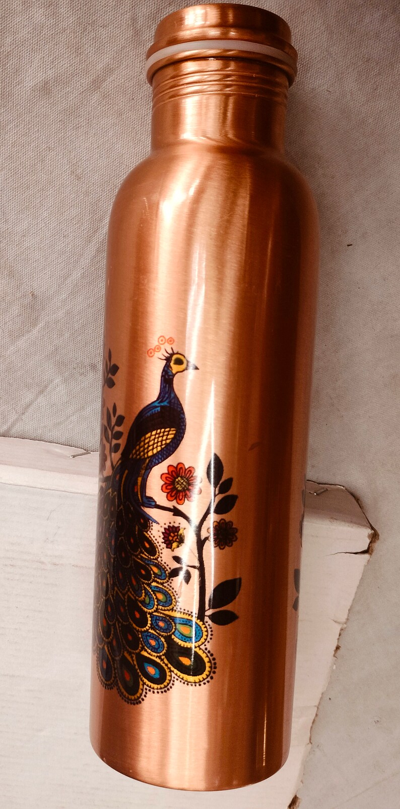 May include: A cylindrical copper water bottle with a metallic finish, featuring a printed graphic of a peacock with blue, yellow, and orange plumage perched on a branch with red and orange flowers. The bottle has a matching screw-top lid.