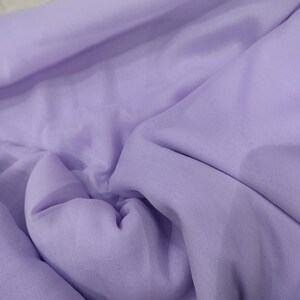 Pode incluir: Close-up de um tecido roxo claro, macio e fluido. O tecido apresenta dobras sutis e sombras que criam profundidade. A delicada cor lavanda sugere um material leve, ideal para vestuário ou decoração.