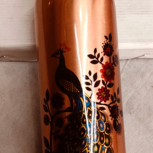 May include: A copper water bottle with a peacock design. The peacock is black, blue, green, and yellow with a colorful tail. The bottle has a copper lid.