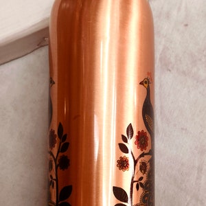 May include: A cylindrical copper water bottle with a polished metallic finish. The exterior features a printed graphic of a peacock perched on a floral branch, with detailed plumage in shades of blue, green, and orange against the copper surface.