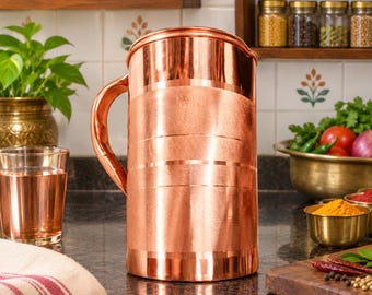 May include: A cylindrical copper pitcher with a polished finish and a series of horizontal engraved lines encircling the body. The pitcher features a sturdy, curved handle on the side and a wide, open top with a small pouring spout.
